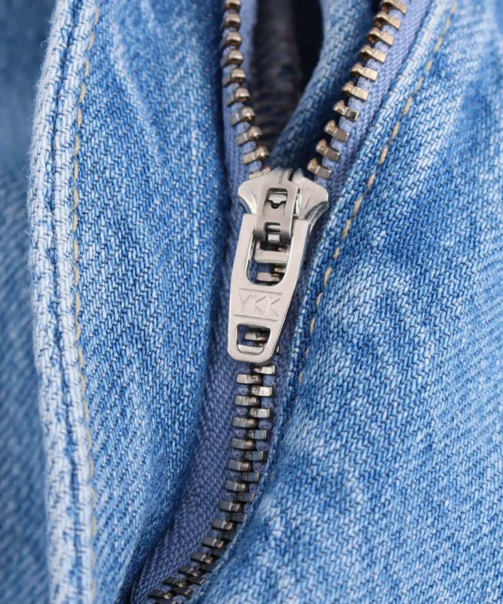 Detalhe macro do zíper frontal com ferragem YKK do macacão jeans pantalona feminino azul claro