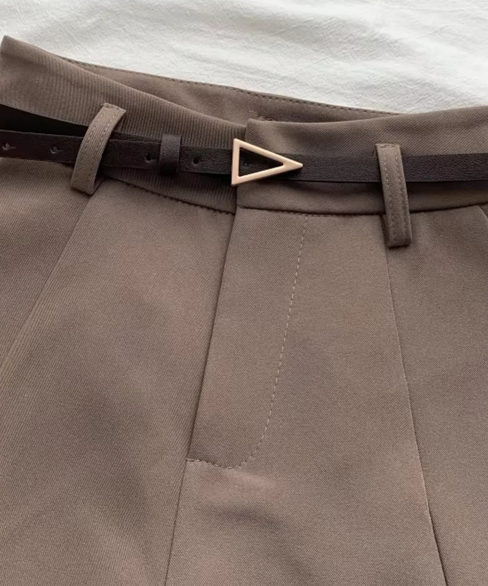 Detalhe do cós do shorts de alfaiataria marrom taupe com o cinto fino escuro passado nas presilhas, fivela dourada e pregas frontais