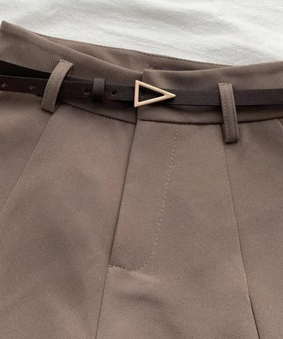 Detalhe do cós do shorts de alfaiataria marrom taupe com o cinto fino escuro passado nas presilhas, fivela dourada e pregas frontais